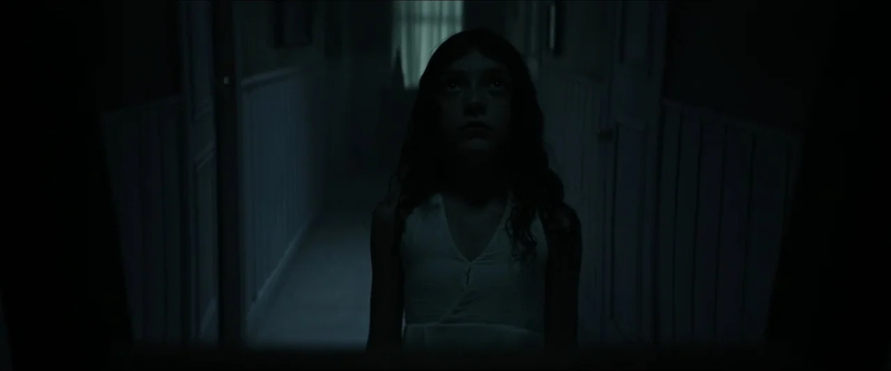 It Feeds (2025) movie still: medium — A dark, narrow residential hallway with closed doors lines the walls on either side. A young girl stands in…