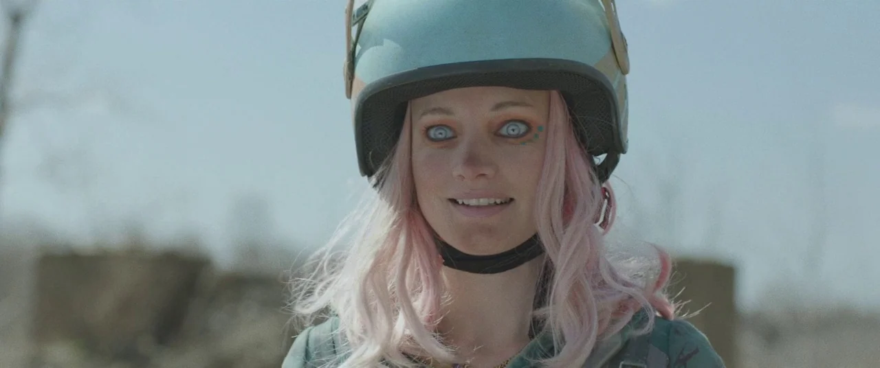 Turbo Kid (2015) movie still: closeup — A woman with pink hair wears a helmet outdoors in a desolate landscape. She looks directly toward the viewer…