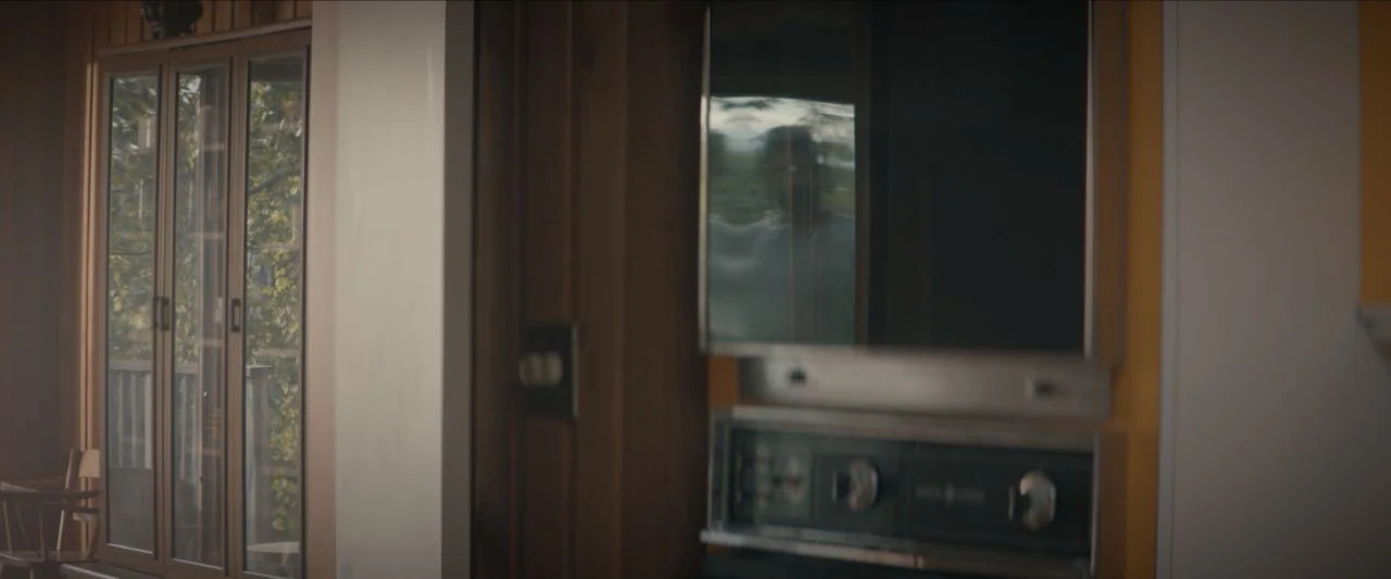 Becky (2020) movie still: medium — A vintage wood-paneled residential space includes a built-in oven with a dark reflective surface. A man is…