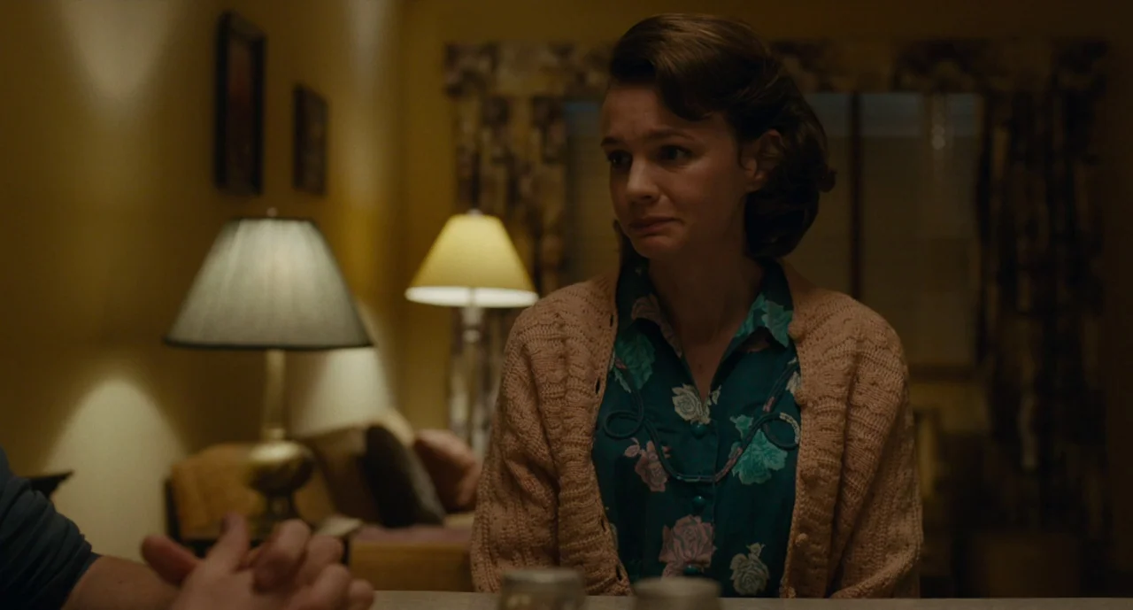 Wildlife (2018) movie still: medium — An interior space features two table lamps casting warm light against a dark wall. A woman wearing a floral…