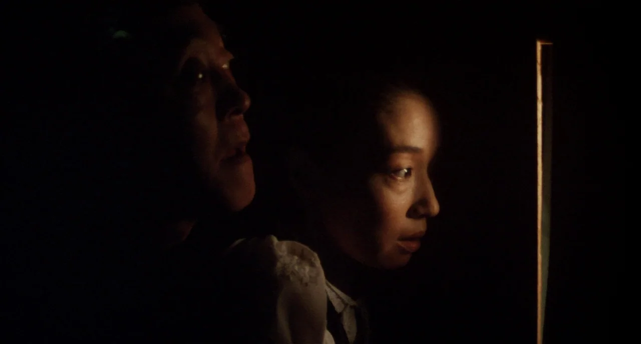 Tampopo (1985) movie still: medium — Two individuals sit in darkness illuminated by a sliver of light. They look toward an unseen source in the dark…