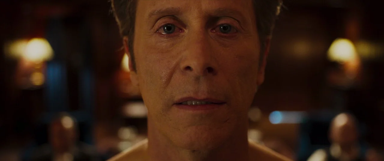 The Perfection (2018) movie still: closeup — A dimly lit room with blurred warm lamps visible in the soft-focus background. A middle-aged man stares…