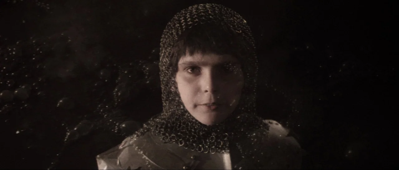Macbeth (1971) movie still: closeup — A person is positioned in a dark, undefined environment. The subject wears a chainmail hood and faces the…