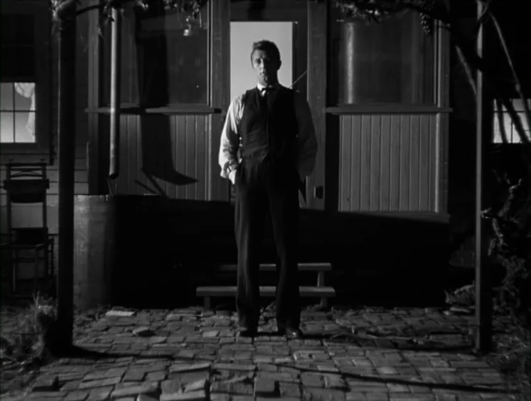 The Night Of The Hunter (1955) movie still: fullbody — A man stands on a brick porch outside a wooden structure at night. He is illuminated by a…
