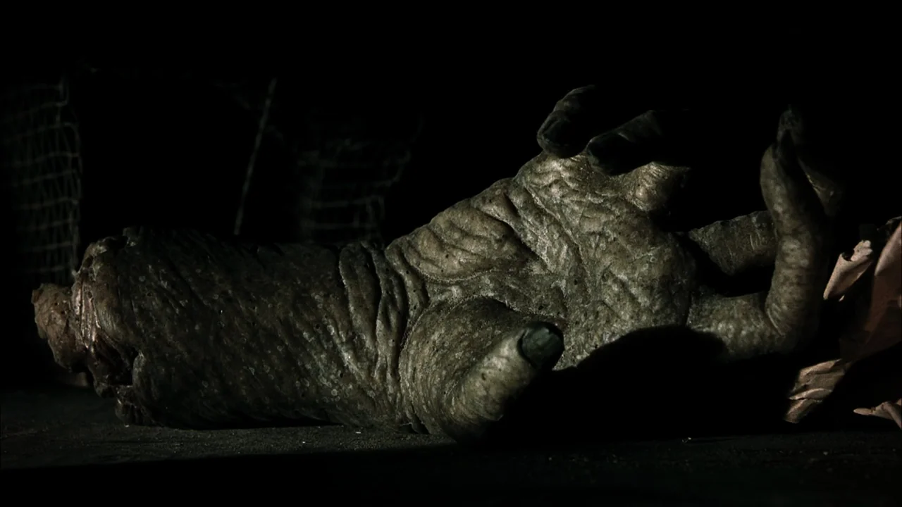 The Texas Chainsaw Massacre (2003) movie still: closeup — A disembodied, textured, grey-green hand lies flat on a dark surface. The surrounding area…