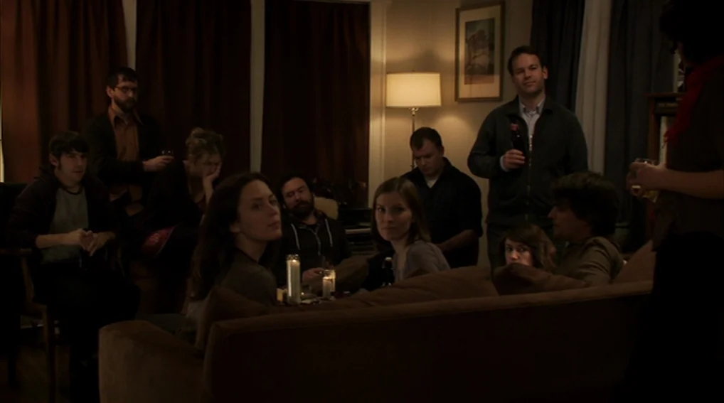 Your Sister’s Sister (2011) movie still: wide — A dimly lit residential living room with several people seated on dark brown sofas. The space has…