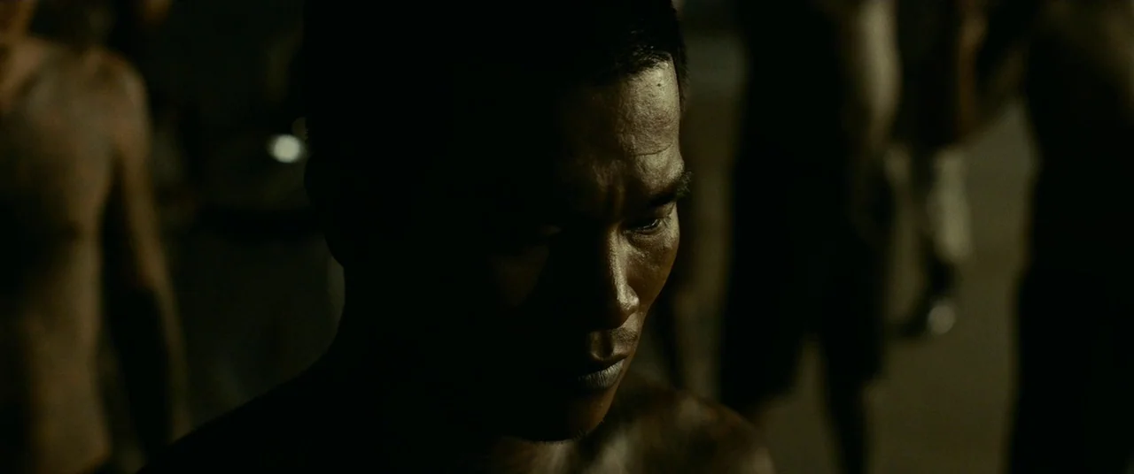A Prayer Before Dawn (2017) movie still: closeup — A dark outdoor space at night with indistinct figures in the blurry background. An shirtless man…
