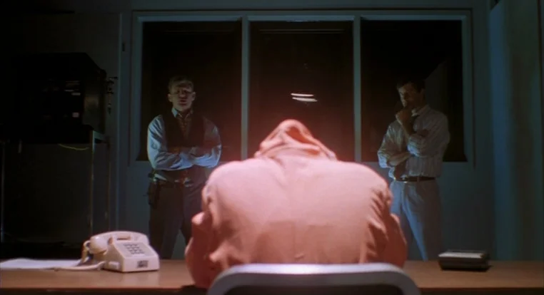 Schizopolis movie still: medium — A dark, spartan room features a desk with a white telephone in the foreground. A person in an orange hood sits…
