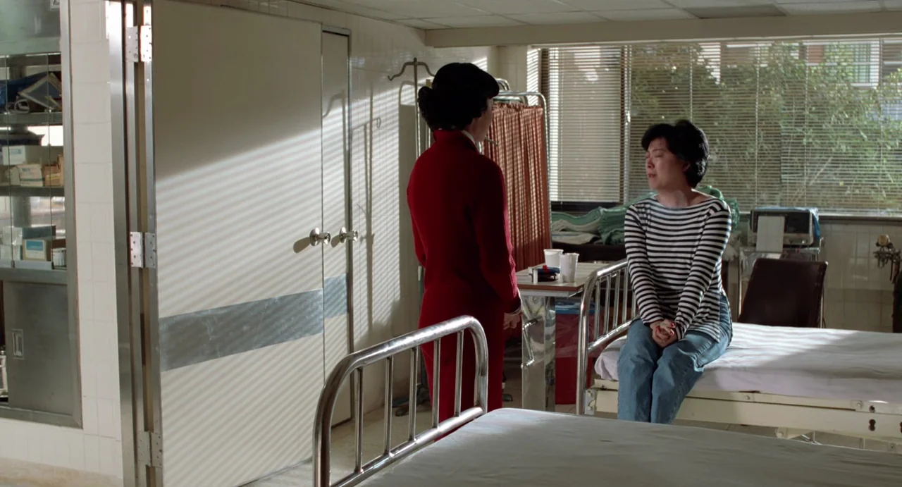 A Confucian Confusion (1994) movie still: medium — A hospital room with metal beds and white tiled walls featuring shuttered window blinds. A woman…