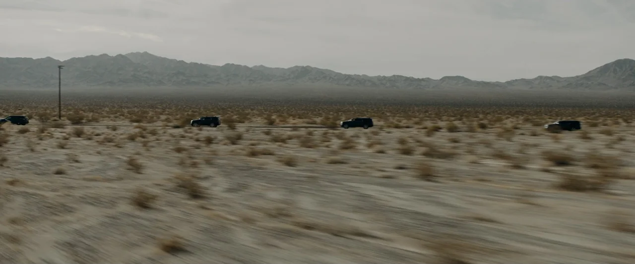 Encounter (2018) movie still: wide — A vast, flat desert landscape stretches toward a mountain range under a gray sky. Four dark-colored SUVs travel…