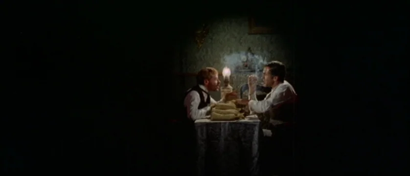 Django Kill (1967) movie still: medium — Two men sit at a small table inside a dimly lit room with wallpapered walls. One man holds a lit cigar…