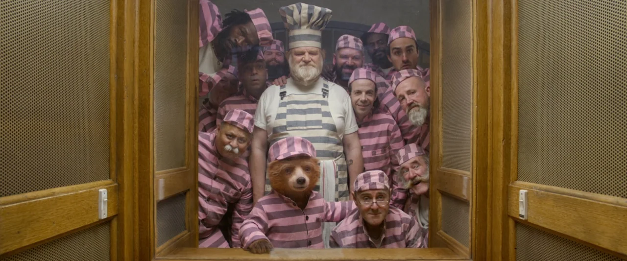 Paddington 2 (2017) movie still: medium — A group of men in striped prison uniforms and a bear pose together inside. They are framed by a doorway…