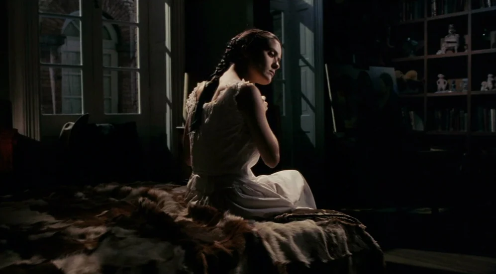 Frida (2002) movie still: medium — A dimly lit room contains a bed covered with a textured brown animal hide. A woman sits on the edge of the bed…