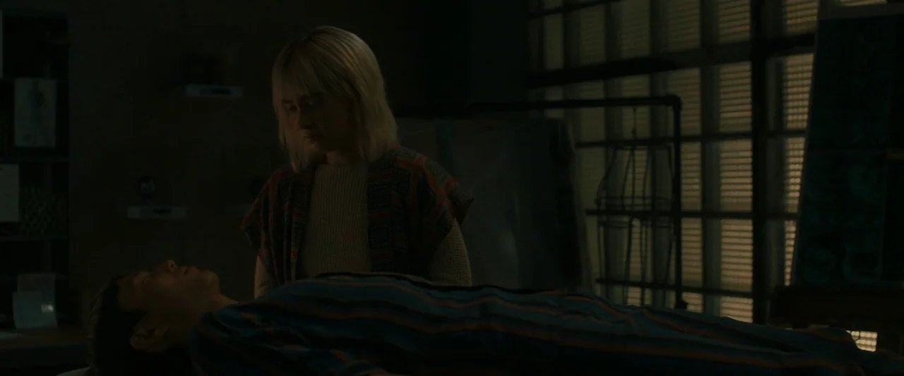 After Yang (2021) movie still: medium — A dimly lit industrial workspace features a glass brick wall in the background. A woman watches over a man…