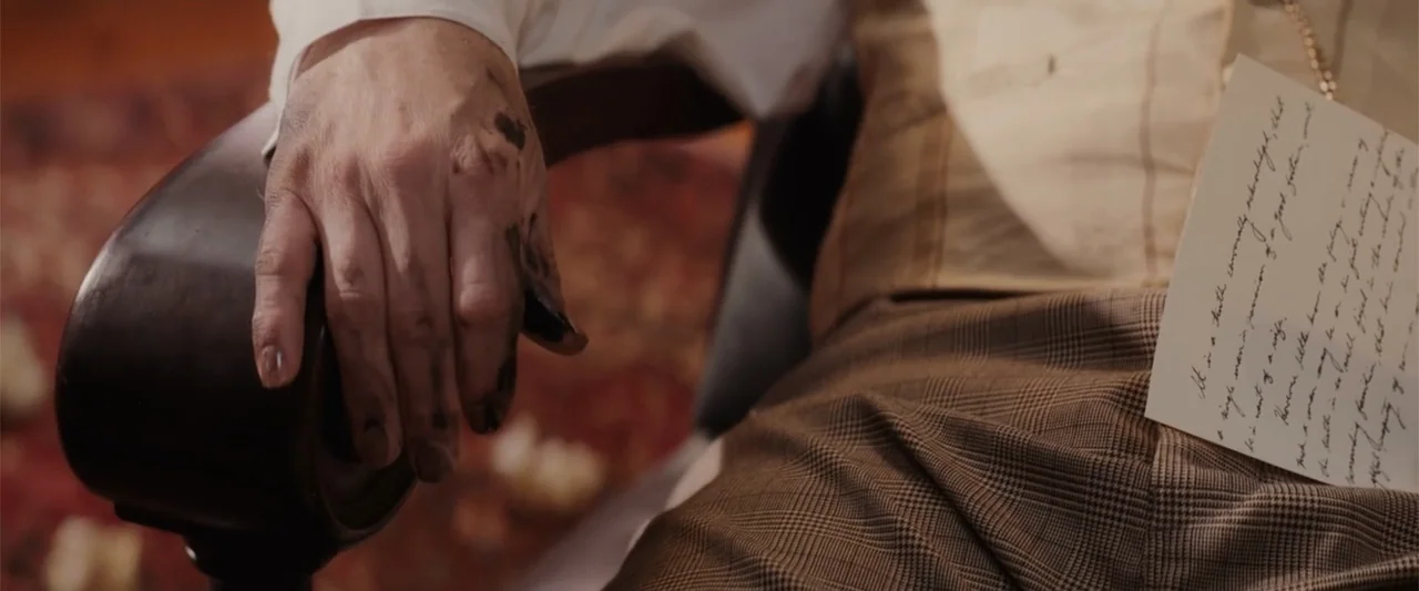 I Heard the Bells (2022) movie still: closeup — An interior space features a person in plaid trousers sitting in a chair. A ink-stained hand rests…