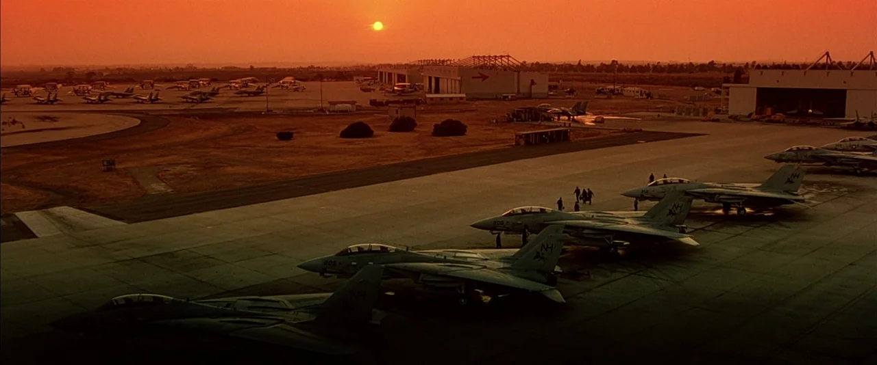 Top Gun (1986) movie still: establishing — A military airbase tarmac with rows of parked F-14 fighter jets at sunset. The orange sky dominates the…