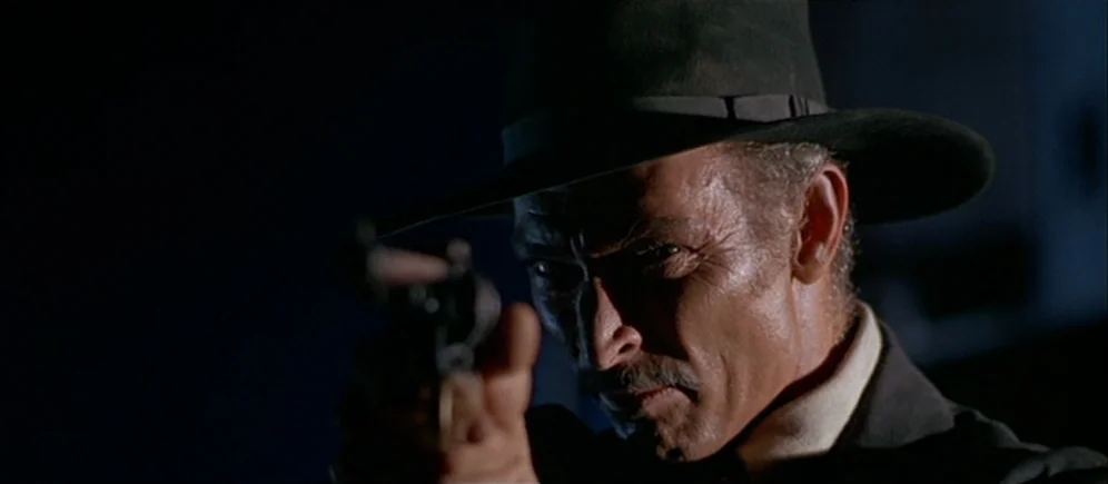 For A Few Dollars More (1965) movie still: closeup — A man stands in extreme darkness with only his face illuminated by sharp light. He holds a…