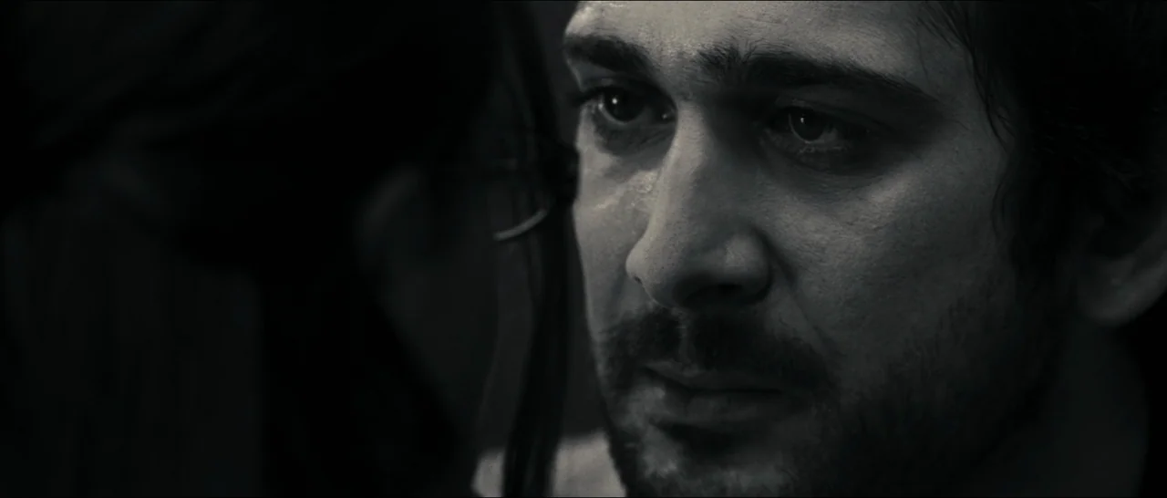Slackers (2002) movie still: closeup — An interior space is dimly lit and shrouded in deep shadow. A man with dark stubble gazes at a person…