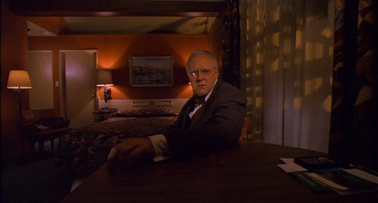 Raising Cain movie still: medium — A dimly lit motel room with terracotta-colored walls and a patterned bedspread. A man in a suit sits at a desk…