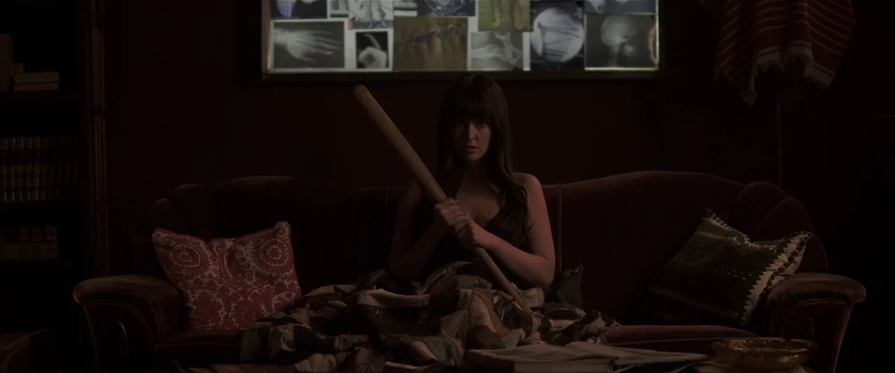American Mary (2012) movie still: medium — A dark living room features a person seated on a velvet couch holding a bat. Behind her, a wall display…