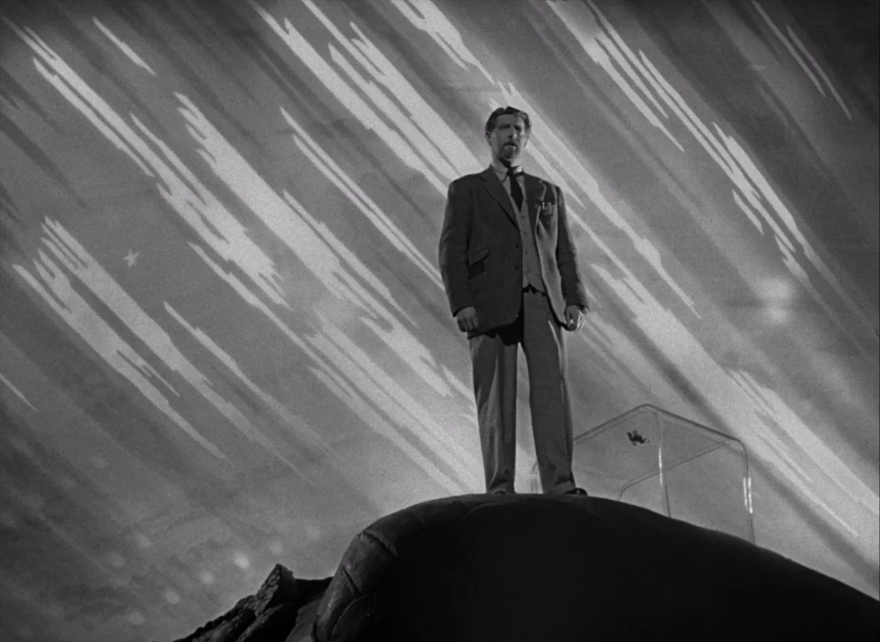 A Matter of Life and Death (1946) movie still: fullbody — A man in a suit stands atop a dark rocky outcropping at night. An stylized painted…