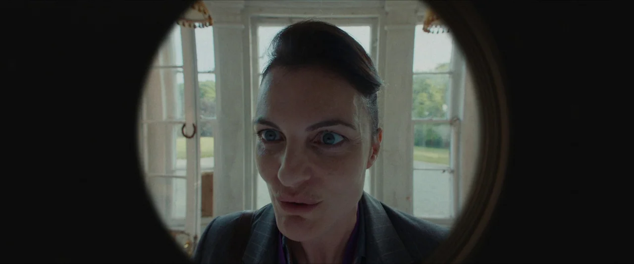 Fréwaka (2024) movie still: closeup — A view through a door peephole showing a woman standing outside a building. She stares intently toward the…