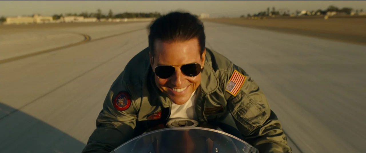 Top Gun: Maverick (2022) movie still: medium — A wide paved runway stretches out under a clear golden hour sky. A man in a flight jacket drives a…