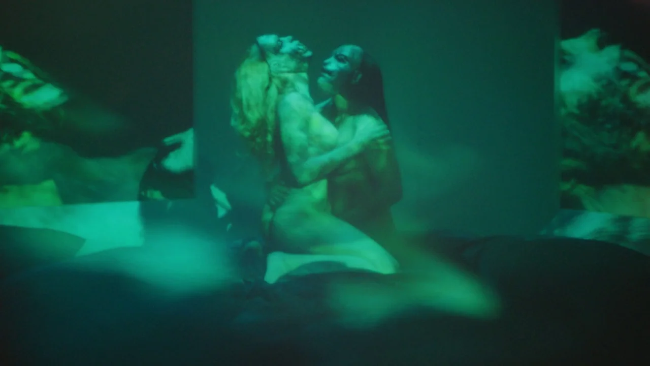 Infinity Pool (2023) movie still: medium — A dark, featureless interior space features a projected image of two figures. The two figures are…