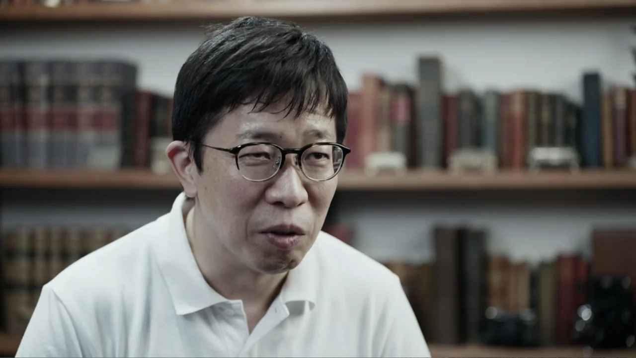 Vai Corinthians (2023) movie still: medium — A man stands before a blurred bookshelf filled with old volumes in a library. He wears a plain white…