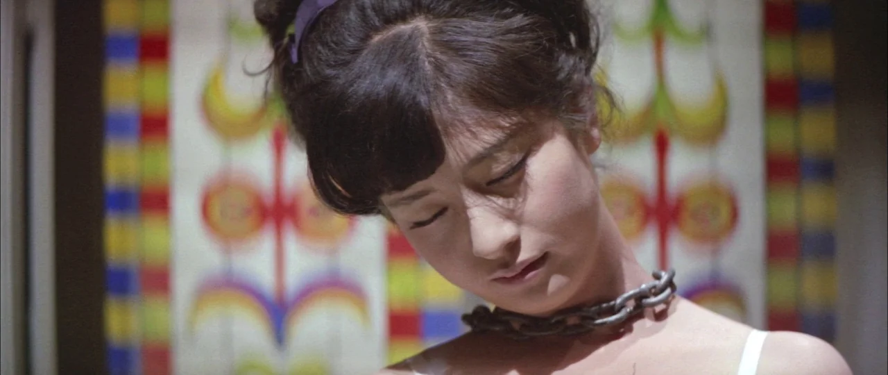 Black Tight Killers (1966) movie still: closeup — A woman stands in front of a white wall adorned with colorful geometric patterns. She wears a…