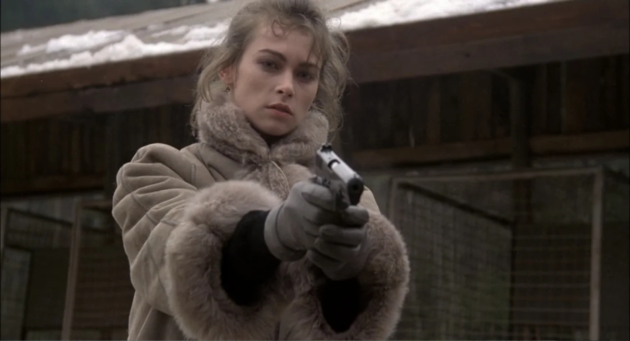 Gorky Park (1983) movie still: medium — A woman stands before a wooden shelter under an overcast sky with scattered snow. She holds a silver handgun…