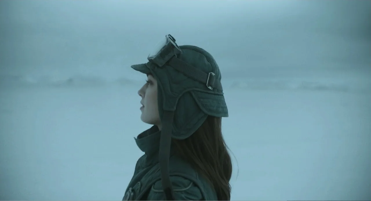 Mickey 17 (2025) movie still: medium — A snowy, overcast plain stretches behind a person dressed in winter aviation gear. A woman in a green cap and…
