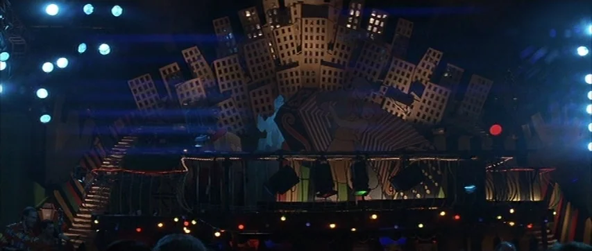 The Truth About Charlie (2002) movie still: wide — An empty stage features a stylized cardboard cityscape backdrop and illuminated circular light…