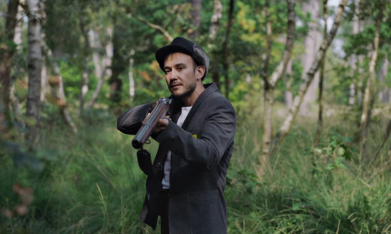 Bloodsuckers (2021) movie still: medium — A dense forest with tall trees and overgrown grass in the foreground. A man stands holding a long gun…