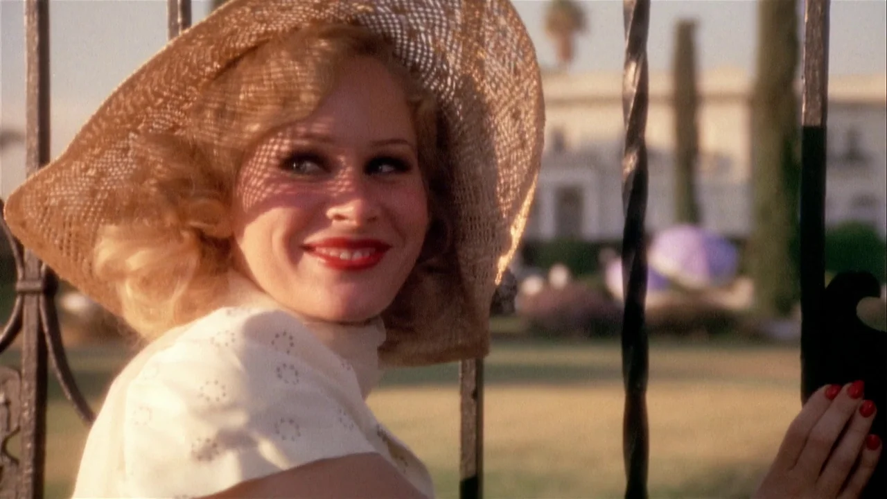 The Day of the Locust (1975) movie still: medium — A woman stands behind a dark metal gate on a sunny estate grounds. She looks over her shoulder…