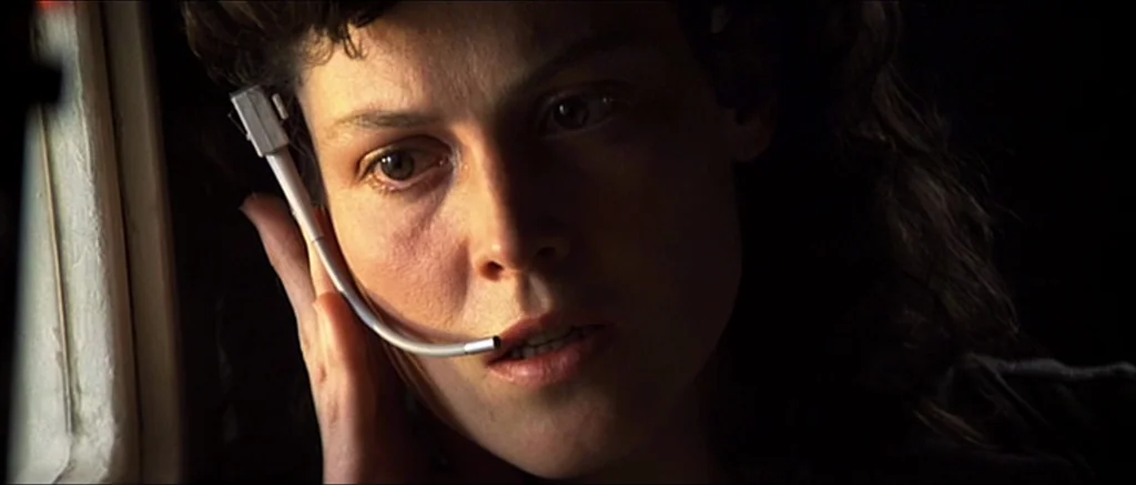 Alien (1979) movie still: closeup — An interior space within a dark, metallic environment with minimal light visible. A woman with dark wavy hair…