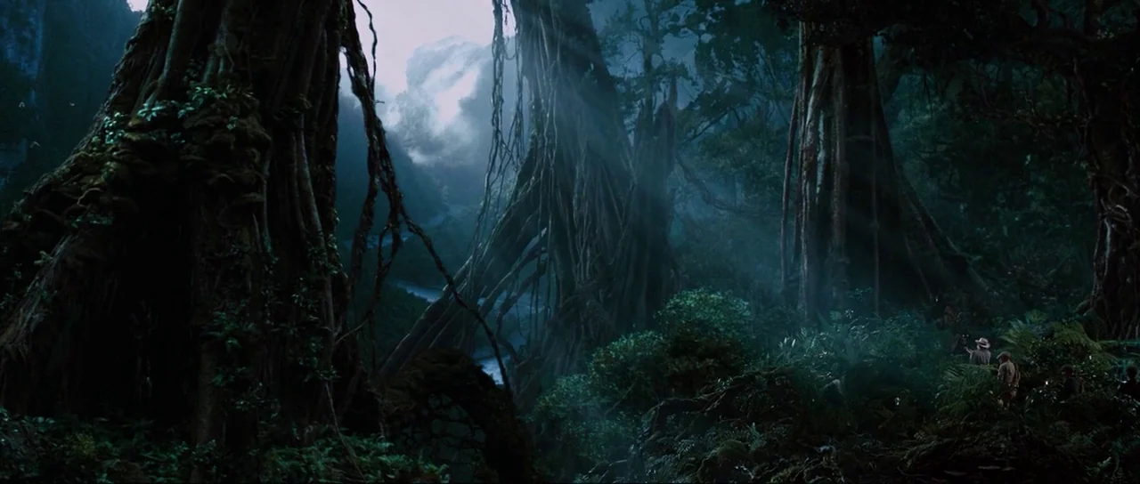 King Kong (2005) movie still: wide — A dense, ancient-looking jungle features enormous trees with thick exposed roots and hanging vines. Light rays…