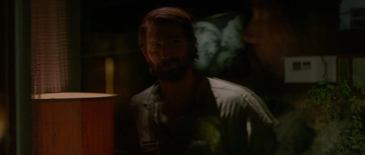 The Invitation (2015) movie still: medium — A man stands in a dimly lit room with blurred architectural features in the background. A table lamp…