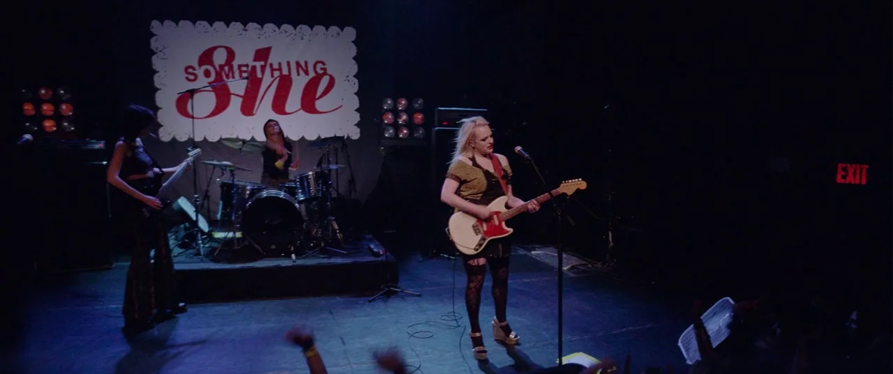 Her Smell (2018) movie still: wide — A dimly lit club stage features a band performing in front of a banner. A female guitarist stands center stage…