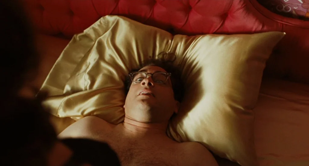A Serious Man (2009) movie still: closeup — A man lies on a bed with gold satin pillowcases and a red headboard. The camera captures him from a high…