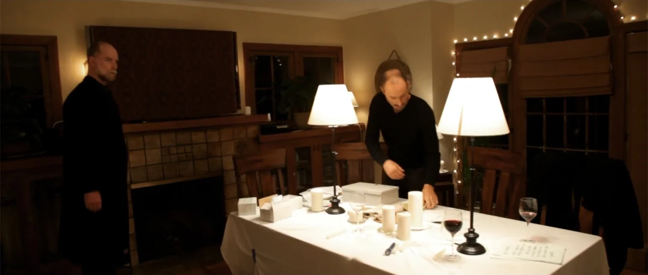 La coherencia del caos movie still: medium — A dimly lit room contains a dining table set with lamps and wine glasses. One man stands by a fireplace…
