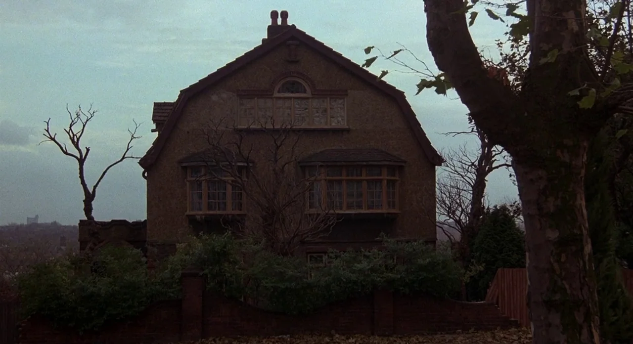 Hellraiser (1987) movie still: establishing — A two-story brown house with prominent bay windows stands behind a low brick wall. Bare, leafless…