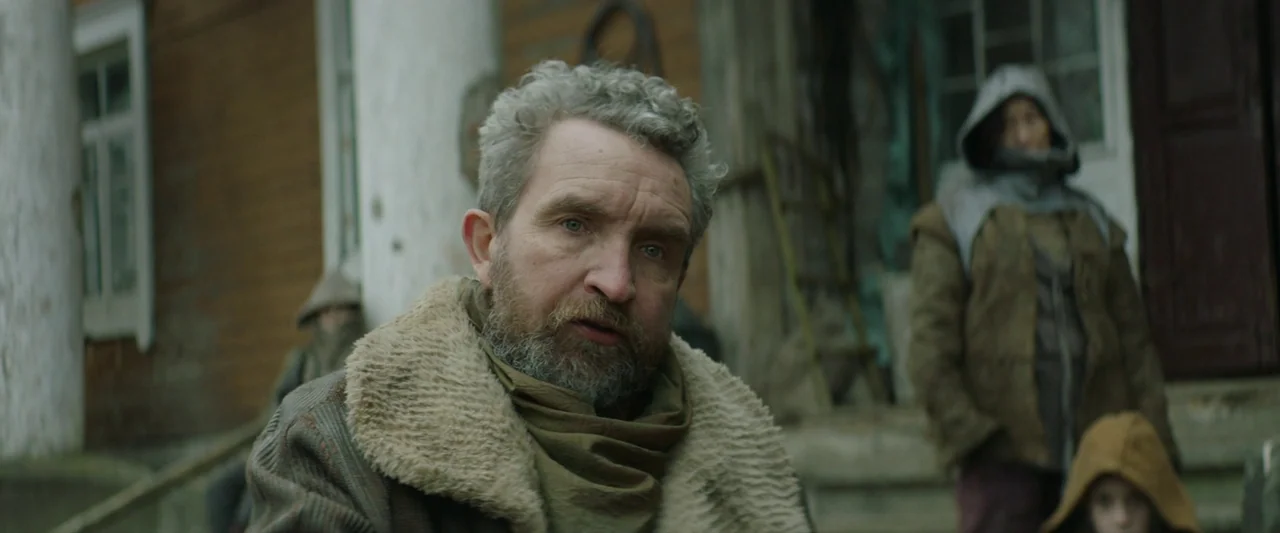 Vesper (2022) movie still: medium — A wooden building with peeling white painted columns serves as the backdrop. The man stands in the foreground…