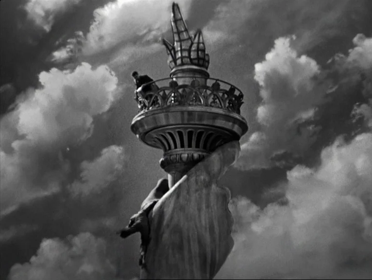 Saboteur (1942) movie still: wide — The massive iron torch and viewing platform of a colossal monument rises toward clouds. A small man stands alone…
