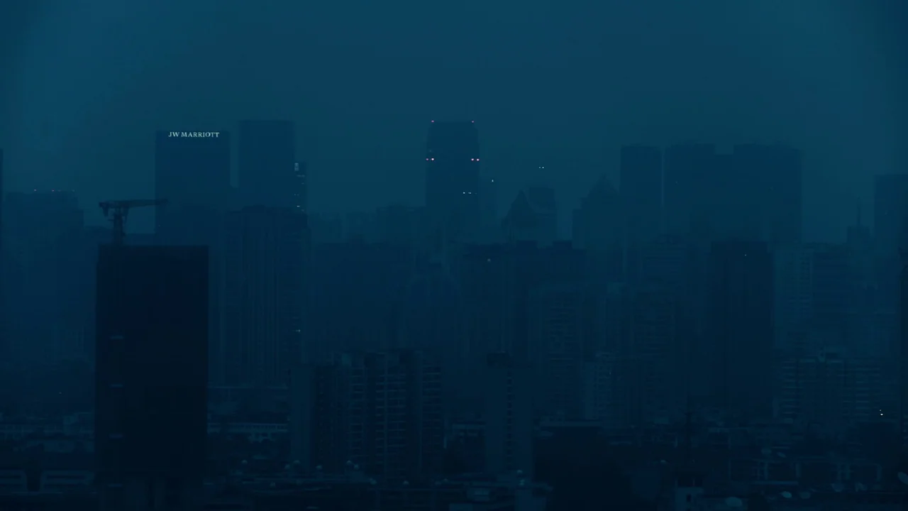 The Last Year of Darkness (2023) movie still: establishing — A dense urban skyline is obscured by heavy atmospheric haze during the blue hour.…