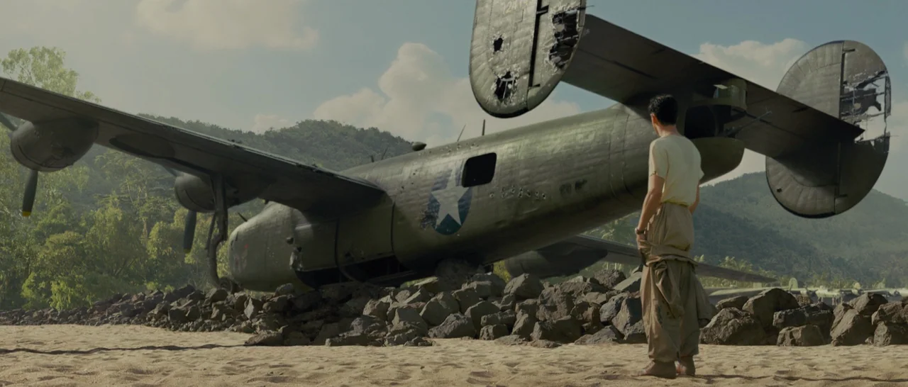 Unbroken (2014) movie still: wide — A damaged green World War II bomber aircraft sits crashed on a sandy beach. A lone man stands with his back to…