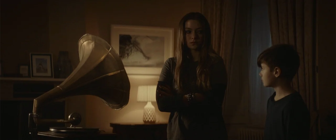 The Cellar (2022) movie still: medium — A dim living room interior features a vintage gramophone and a lit table lamp. A young woman and a young boy…