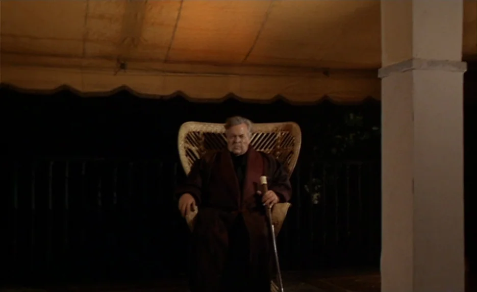 The Immortal Story (1968) movie still: medium — A man sits on a dark veranda at night under a low canvas roof. He sits in a wicker chair holding a…