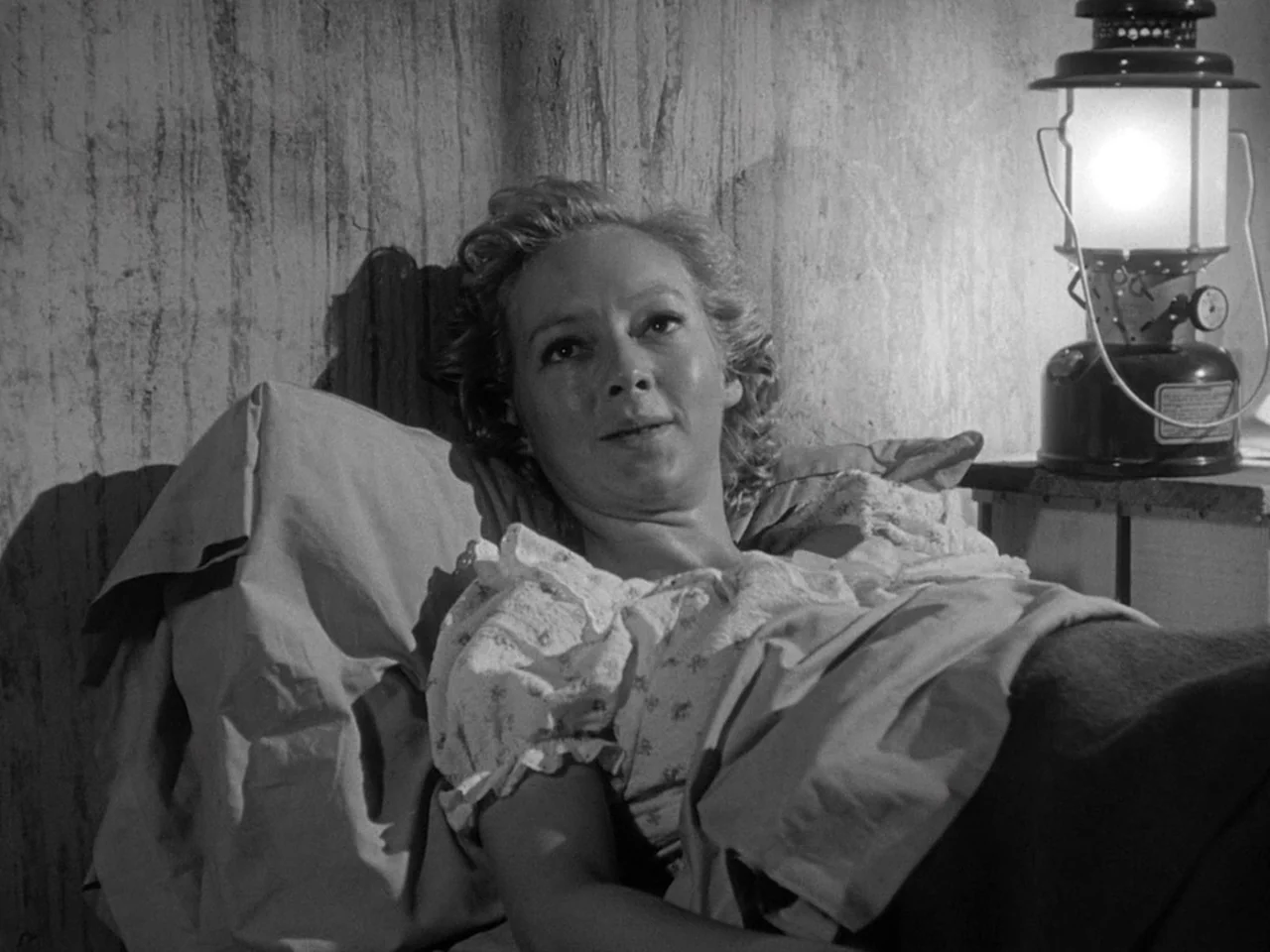 The Prowler (1951) movie still: medium — A woman lies against pillows in a room with textured, worn walls. A metal lantern sits on a surface near…