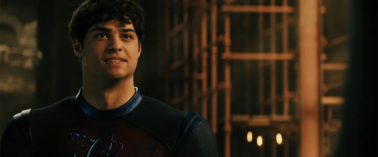Black Adam (2022) movie still: medium — A dimly lit room with blurred industrial scaffolding visible in the background. A young man in a…